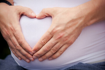 husband hugs his pregnant wife by the stomach and makes fingers in the shape of a heart.