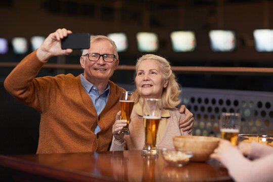 Portrait of modern senior couple taking selfie photo while drinking beer in bar and enjoying night out with friends, copy space