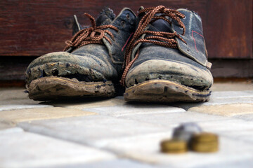 Worn boots with a lagging sole on a background of gray pavers and dark brown wood. In the foreground is a blurred stack of coins. The concept of poverty, homelessness, lack of money. Selective focus