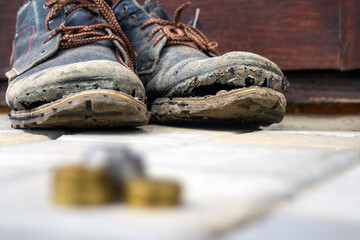 Worn boots with a lagging sole on a background of gray pavers and dark brown wood. In the foreground is a blurred stack of coins. The concept of poverty, homelessness, lack of money. Selective focus. 