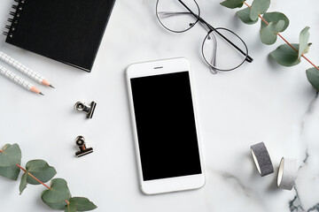Modern feminine workspace with smartphone mockup, glasses, eucalyptus branches, notebook and office supplies on marble desk. Flat lay, top view. Elegant style.