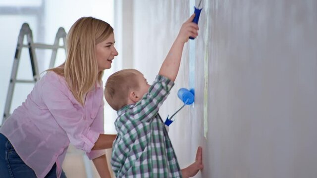 game with disabled child, young smiling woman has fun with boy with down syndrome and paints walls with paint using rollers indoors during repairs