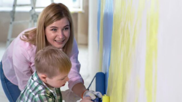 entertainment children, young female parent helps son with down syndrome paint walls with construction roller with colored paints indoors during renovation