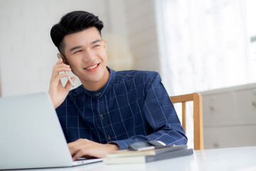 Young asian man talking phone and work from home with laptop computer, freelance using notebook to internet online and speak on smartphone, domestic life, business and communication concept.