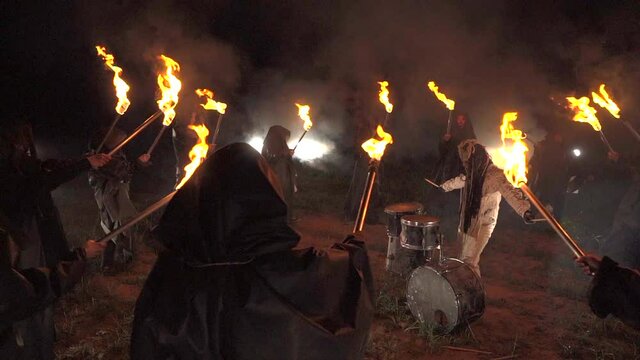 A drummer plays drums in a field at night. Around people with torches.