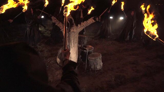 A drummer plays drums in a field at night. Around people with torches.