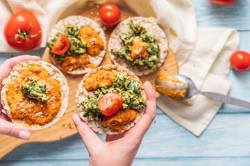 Crispy puffed rice cakes on table with hummus spread spinach with eggs and tomato and vegetables on the table - top view hands holding on healthy vegetarian breakfast gluten free - bright filter
