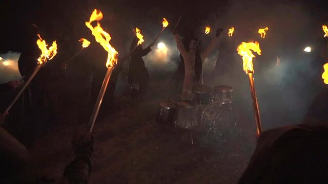 A drummer plays drums in a field at night. Around people with torches.