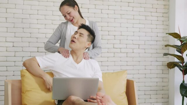 Happy Asian family couple husband and wife laughing sitting on sofa using laptop computer working from home. Business man sitting on sofa home working on laptop her woman is giving her shoulder massag