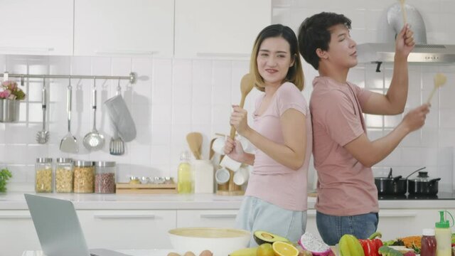 Happy Asian beautiful family couple husband and wife singing in kitchenware microphones in kitchen together having fun dance listen music at home. Two people dancing