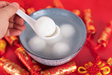 A bowl of Tangyuan or Yuanxiao surrounded by red firecrackers on the red background and a hand-held spoon scooping a tangyuan