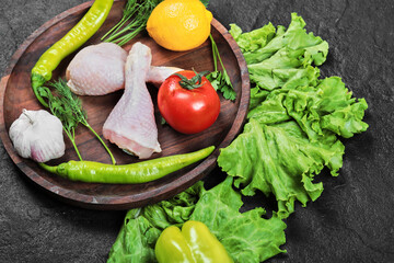 Raw chicken legs on wooden plate with greens, tomato, garlic, peppers and lemon