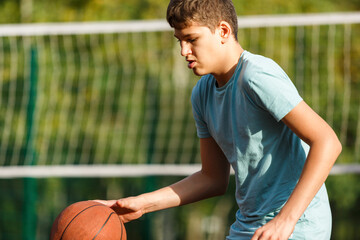 Cute young boy plays basketball on street playground in summer. Teenager in green t shirt with orange basketball ball outside. Hobby, active lifestyle, sport activity for kids.