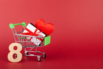 Red heart, gift box with red ribbon inside mini grocery cart on colored background