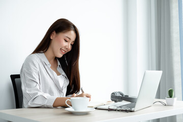 Asian female college students study online at home, enjoy extra lessons, watch videos, and take notes from a digital laptop, do homework by looking at the screen while sitting at a desk at home