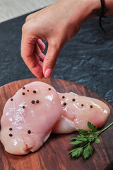 Woman hand adds grain peppers to plate of raw chicken fillet on dark table