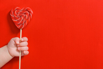 Newborn baby hand holding sweet candy heart on stick on red background