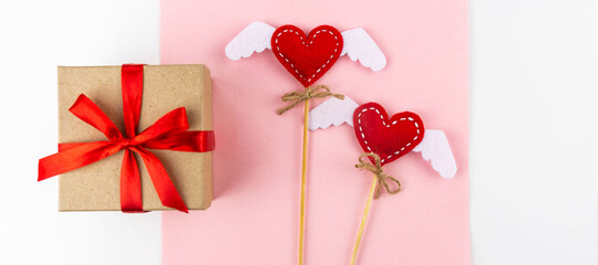 Background Is Valentine's Day. Gift with red ribbon, two hearts with wings on handmade sticks on pink white-paper white background view from above.