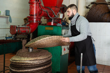 Confident male worker forming pile from fiber mats with milled olive paste, preparing to press on artisanal olive oil factory..