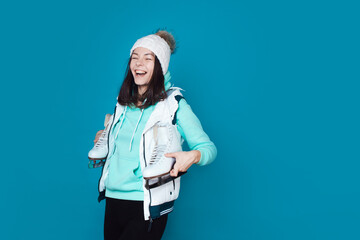 Winter hobby, ice skating. A brunette woman in a hoodie with figure skating skates on her shoulder. Studio shot on a blue background