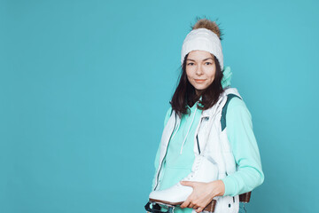 Winter hobby, ice skating. A brunette woman in a hoodie with figure skating skates on her shoulder. Studio shot on a blue background