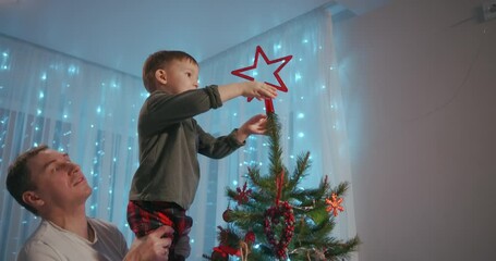 Dad lifted the boy to the top of the Christmas tree to place a Christmas star on top of the top. Father and son decorate Christmas tree together