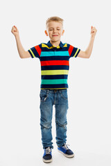 A blond boy in jeans and a multicolored T-shirt stands with his eyes closed and his hands up. White background. Vertical.