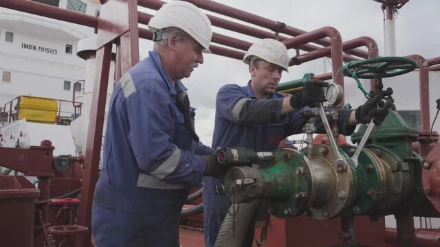professional workers in helmets and jumpsuits repair large pipe with silver spanners under red pipelines against cloudy sky