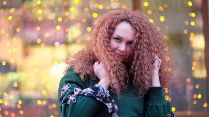 cheerful woman is touching her curly red hair, fooling around against the background of glowing bulbs.