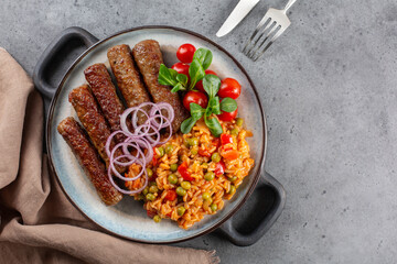 Djuvec rice cooked with red bell pepper, tomato, peas, paprik and beef cevapcici. Grey background,...