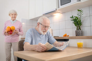 Senior grey-haired man sitting at the table in the kitchen and reading a newspaper