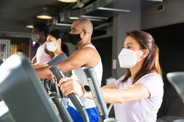 Fototapeta premium Active people in protective masks having running elliptical trainer class in health club