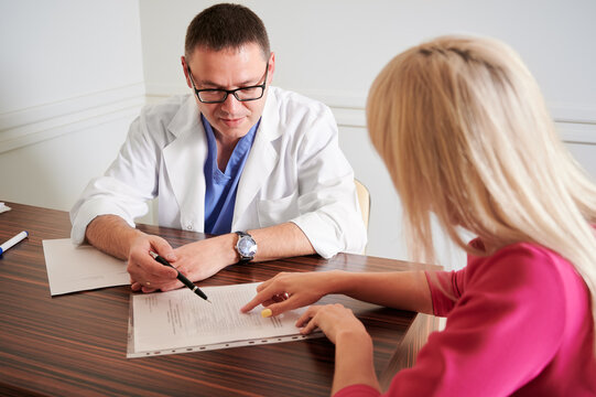 Male Surgeon Talking With Female Patient In Office. Blonde Young Woman Discussing Plastic Surgery With Doctor In Modern Clinic. Concept Of Medical Appointment And Cosmetic Surgery.