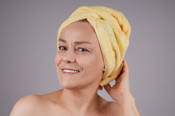 A mature woman with bare shoulders with a towel on her head smiling looks at the camera. Skin care concept. Isolate on gray background