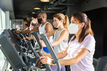 Active people in protective masks having running elliptical trainer class in health club