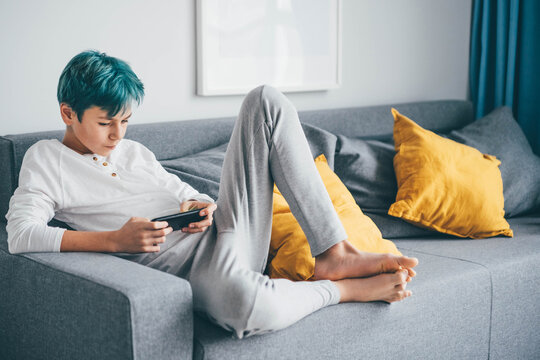 Teenager Using Mobile Phone On The Sofa At Home. Teen With Blue Hair Looks At Phone.