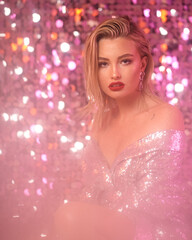 Fashionable partly defocused blurred studio portrait of young sexy blonde woman with makeup and middle length straight hair in shining silver dress against glamor sequins background. Pink light