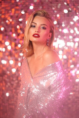 Fashionable partly defocused blurred studio portrait of young sexy blonde woman with makeup and middle length straight hair in shining silver dress against glamor sequins background. Pink light