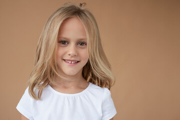 Closeup front portrait of pretty little girl with wavy blond hair, dressed in white shirt, over beige background.