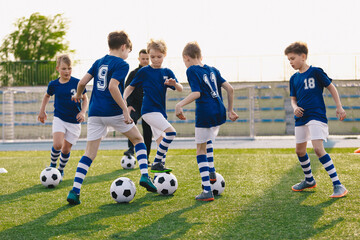 Obraz na płótnie Canvas Group of children playing soccer on training session. Kids in football club wearing blue jersey shirts and soccer kits. Happy boys practicing football with coach on a sunny day