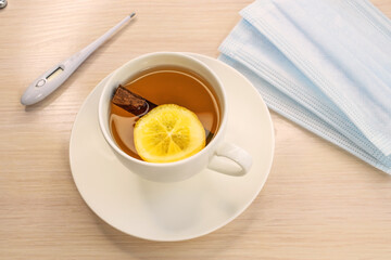 A white cup of lemon tea sits on a wooden table. There is a thermometer and a medical mask near the cup. The concept of prevention and treatment of coronaviruses and influenza viruses