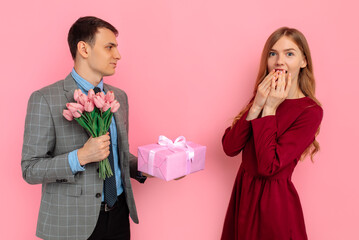 Handsome elegant man giving a present to his beautiful girlfriend over pink background, Valentine's Day concept