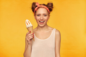 Teenage girl, happy looking red hair woman with two buns. Wearing white tank top and red doted hairband. Holding ice cream and have a big smile. Watching at the camera isolated over yellow background