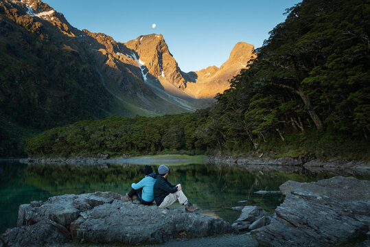 A couple sitting on the rocks and enjoying the views of Lake Mackenzie at Routeburn Track in South Island. Moonrise and sunset at the same time.
