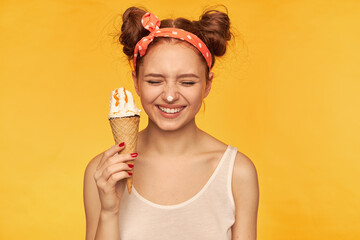 Young lady, pretty ginger woman with two buns. Wearing white tank top and red doted hairband. Holding ice cream and have it on her nose, smiling. Stand isolated over yellow background