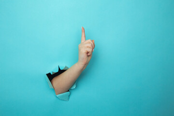 Woman hand breaking through blue paper wall with finger point up.