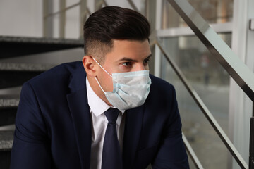 Sad man in protective mask on stairs indoors. Self-isolation during coronavirus pandemic