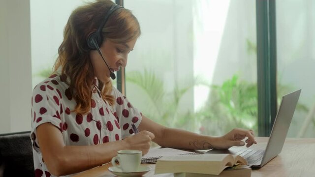 Female call center worker sitting desk focus on customer service member girl in headset looks at laptop screen write in notebook talk with client provide insurance information sell company product