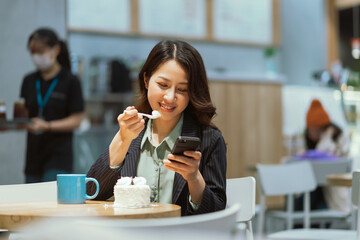Portrait of young Asian businesswoman drinking coffee, eating cream cake and using phone