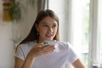 Voice dialing. Smiling young lady hold modern mobile phone in hand use speakerphone or dictate...